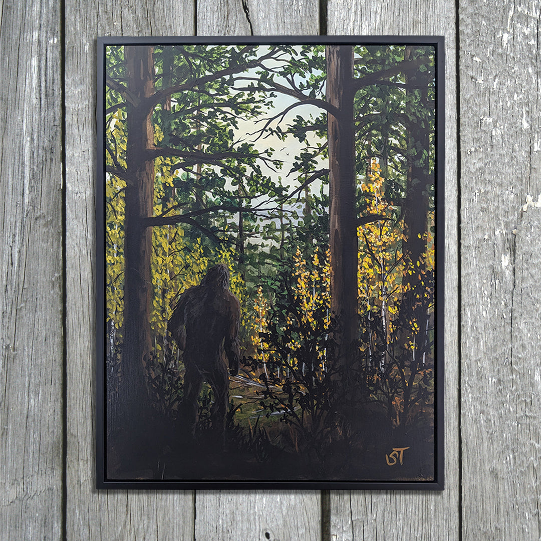 Framed original Sasquatch painting displayed in a black frame against weathered gray wooden boards, showing a shadowed figure moving through a forest of tall trees and autumn leaves, displayed against a wooden background.