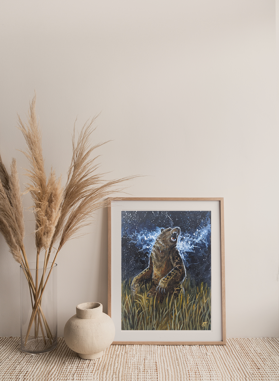 A framed art print depicting a stylized bear sitting in a grassy area with a starry sky in the background, placed on a shelf next to a vase with dried plants.