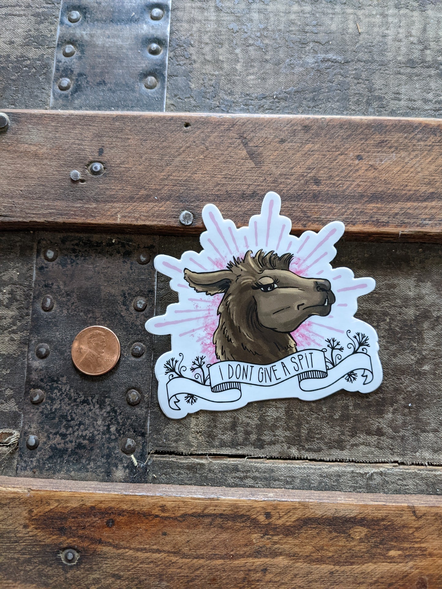 Llama vinyl sticker with the text “I don’t give a spit,” shown next to a penny for size reference on a wooden surface.