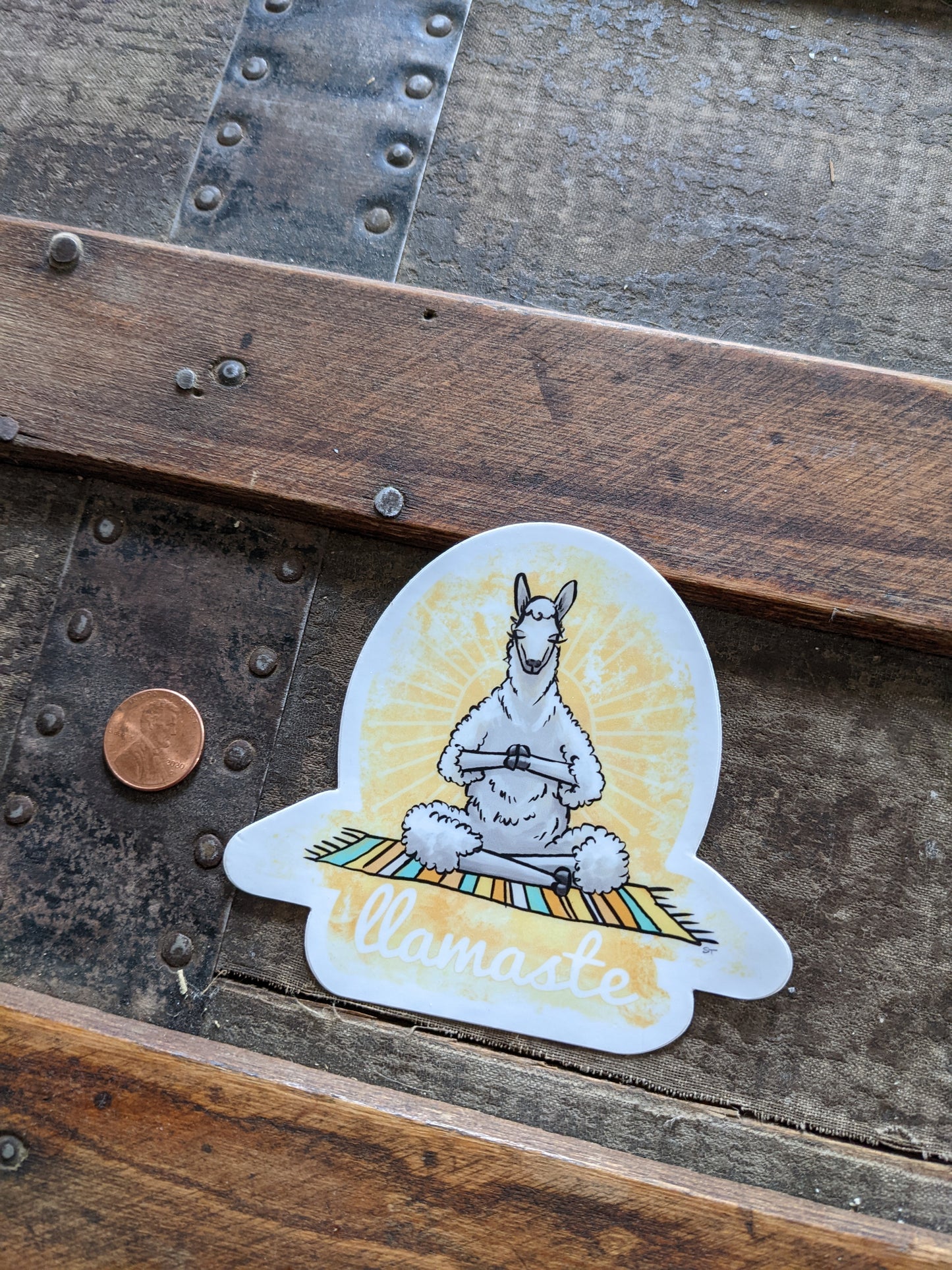 Meditating llama “llamaste” vinyl sticker shown next to a penny for size reference on a wooden surface.