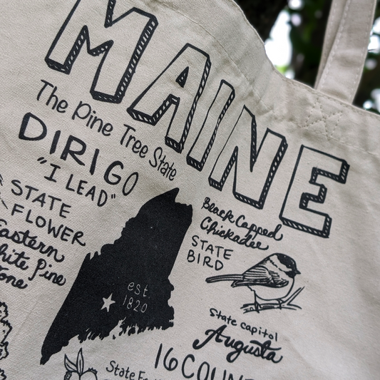 Close-up detail of the Maine-themed tote bag showing illustrated text, the state outline, and chickadee artwork.