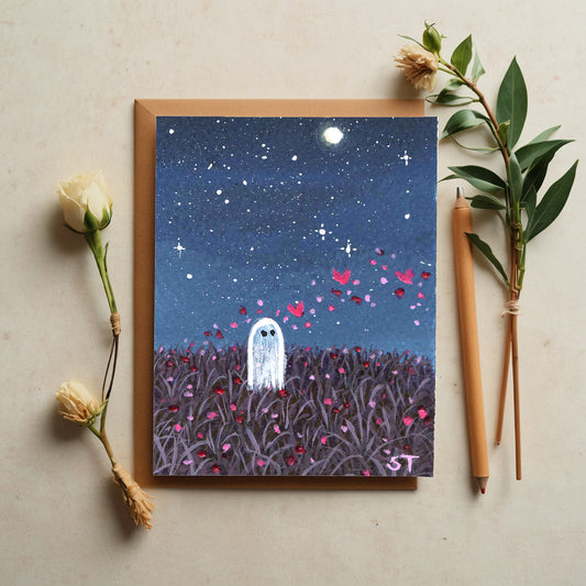 Greeting card with ghost and starry night design, surrounded by flowers and pencils on a beige surface.