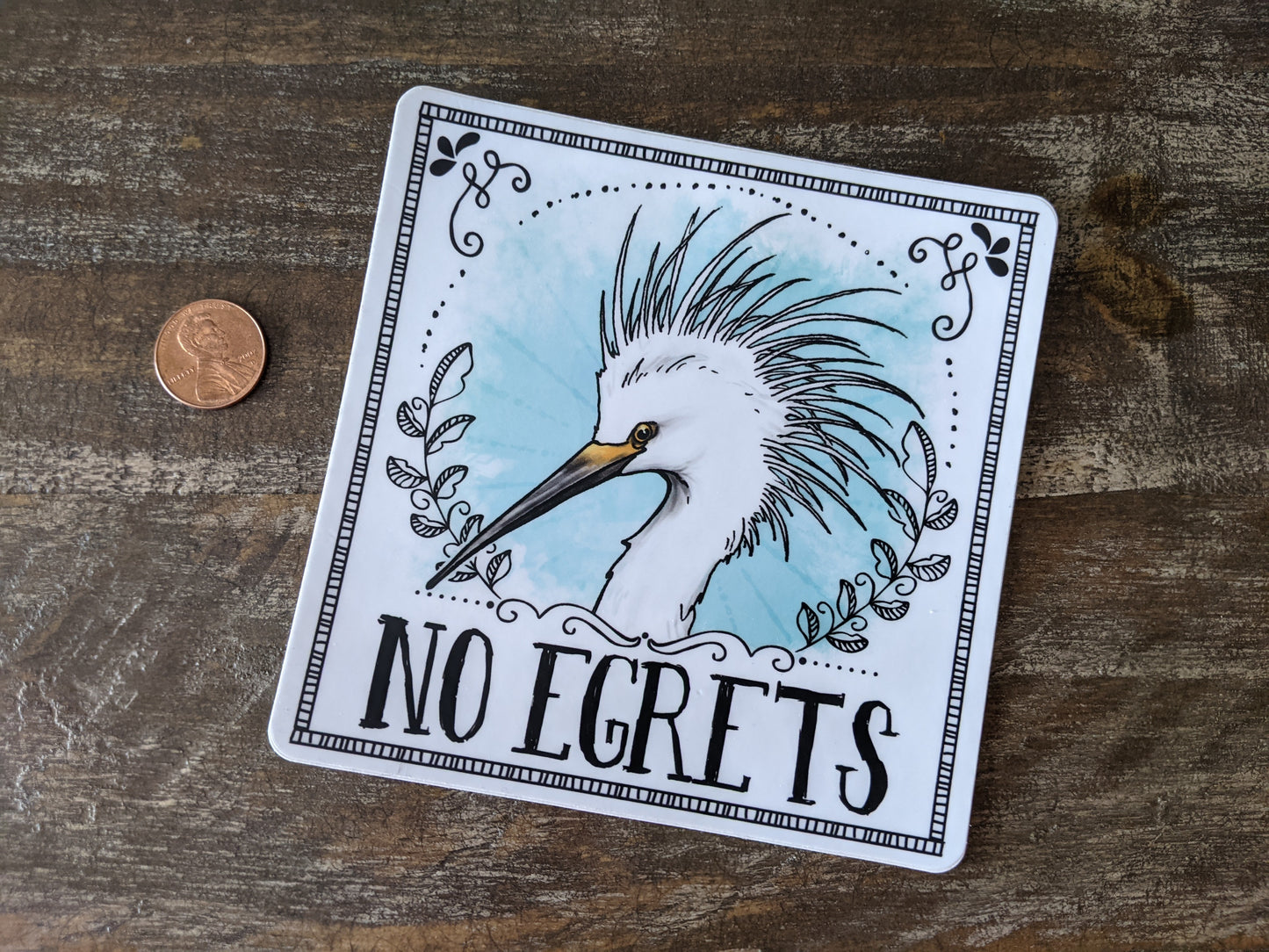 Egret vinyl sticker with decorative framing and the text “No egrets,” shown next to a penny for size reference on a wooden surface.
