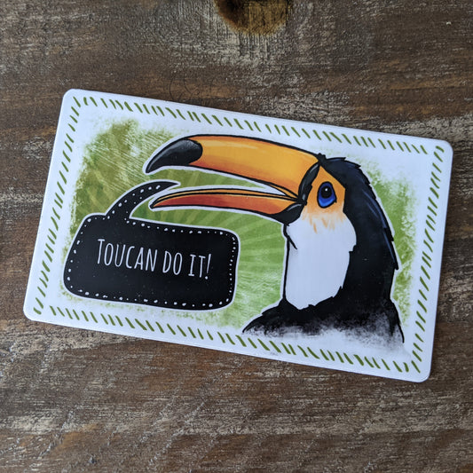 A vinyl decal featuring a hand-illustrated toucan with the text 'Toucan do it!' written in a speech bubble, placed on a wooden surface.