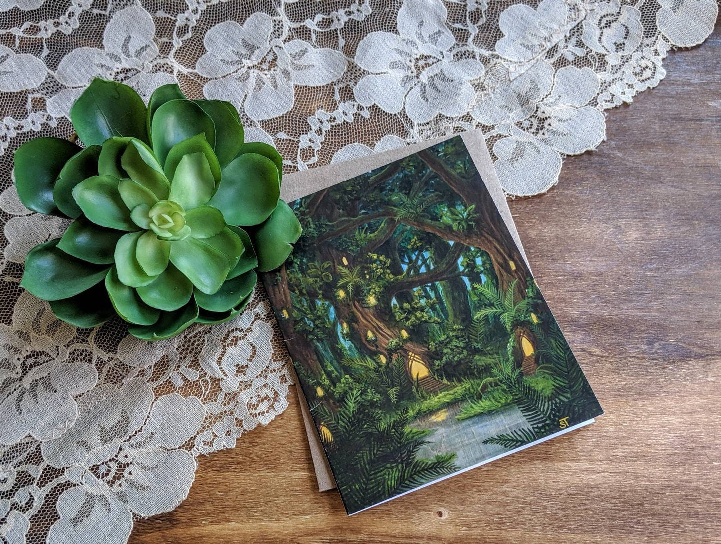 Greeting card showing a lush fantasy forest with glowing fairy house lights among towering trees, styled on a wooden surface with lace fabric and a green succulent.
