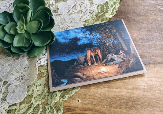 A greeting card with an acrylic painting of a cowboy and his horse near a campfire in the mountains, displayed on a wooden surface with a lacey background.