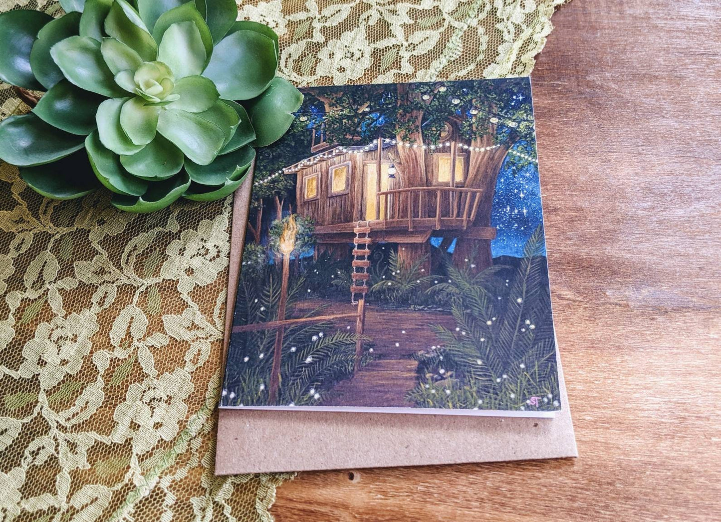 Greeting card showing a warmly lit treehouse at night surrounded by lush greenery and fireflies, styled on a wooden surface with lace fabric and a green succulent.