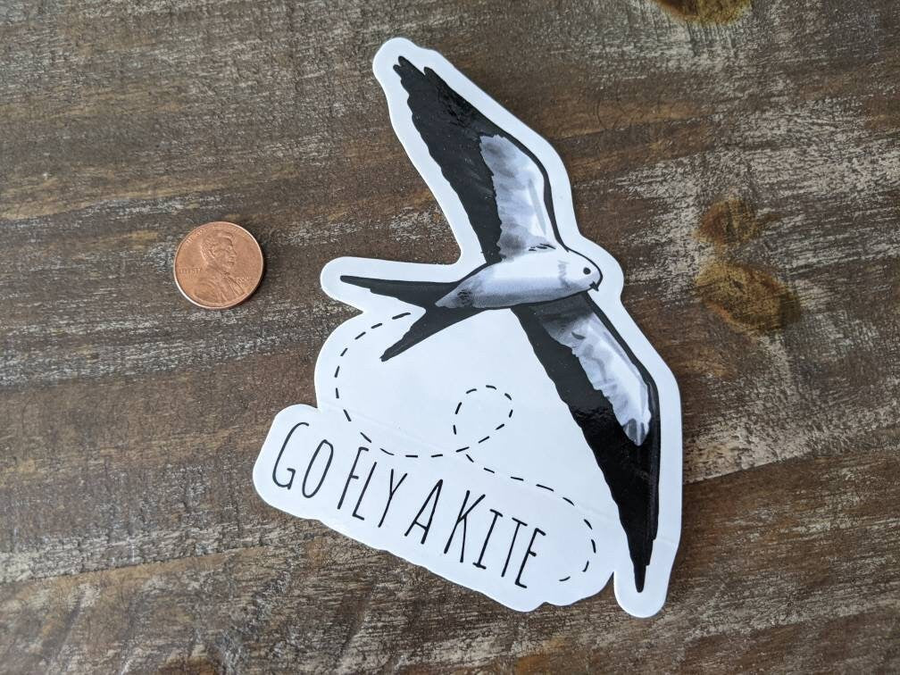 A decorative sticker with a Kite (Bird) illustration and the phrase 'GO FLY A KITE' on it. It appears to be made of glossy adhesive vinyl and is placed on a wooden surface next to a penny for scale.