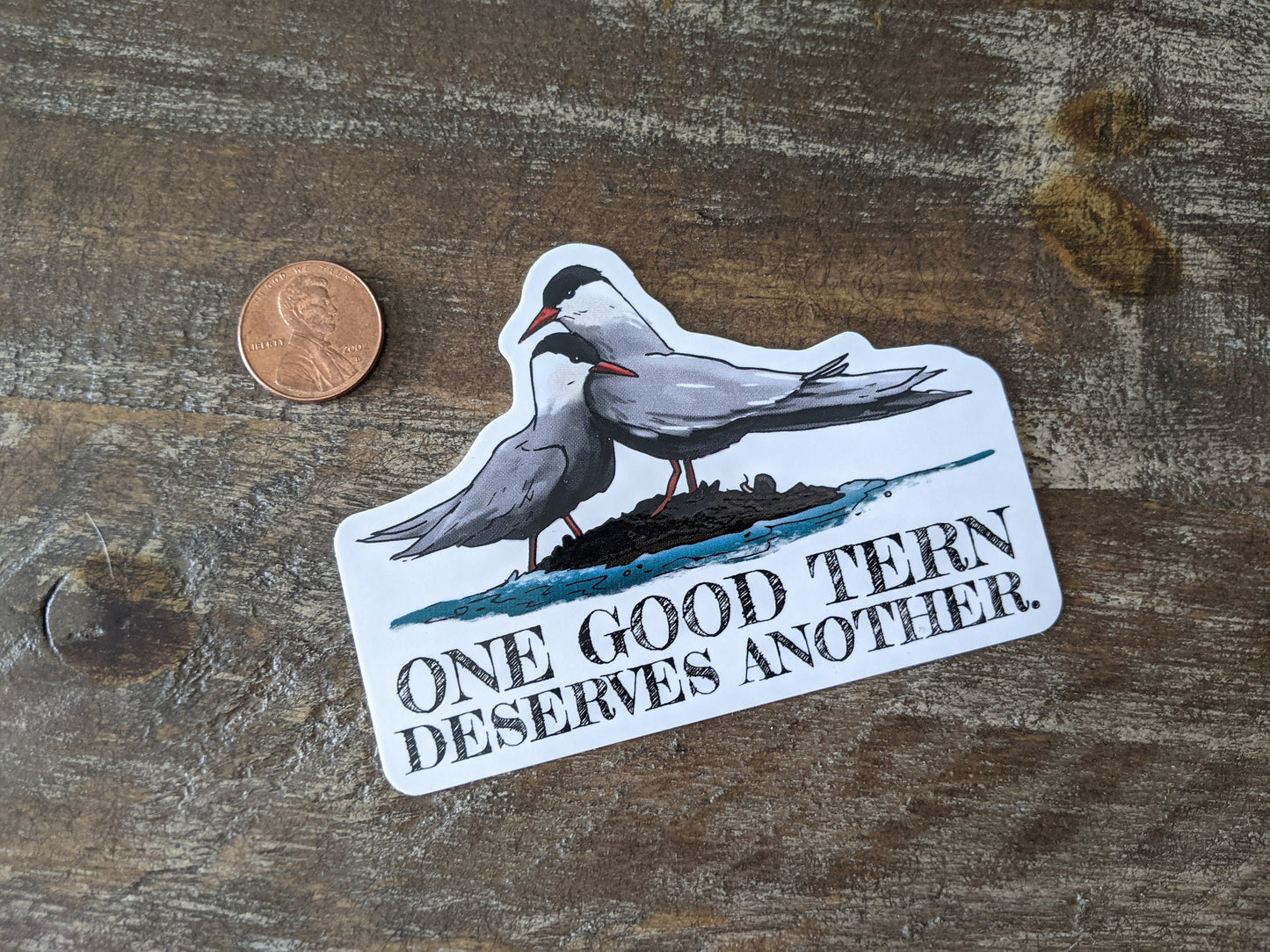 A vinyl decal with a hand-illustrated image of two tern birds and the pun 'one good Tern deserves another' written on it, placed on a wooden surface next to a penny for scale.