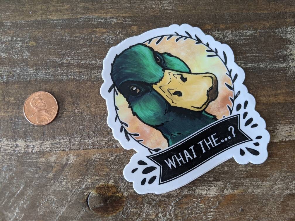 Duck vinyl sticker with a banner reading “What the…?” shown next to a penny for size reference on a wooden surface.