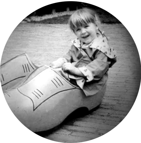 Black and white circular photo of the smiling young child, Sarah Terrell, sitting in a giant clog shoe, outdoors, conveying a playful, nostalgic moment.