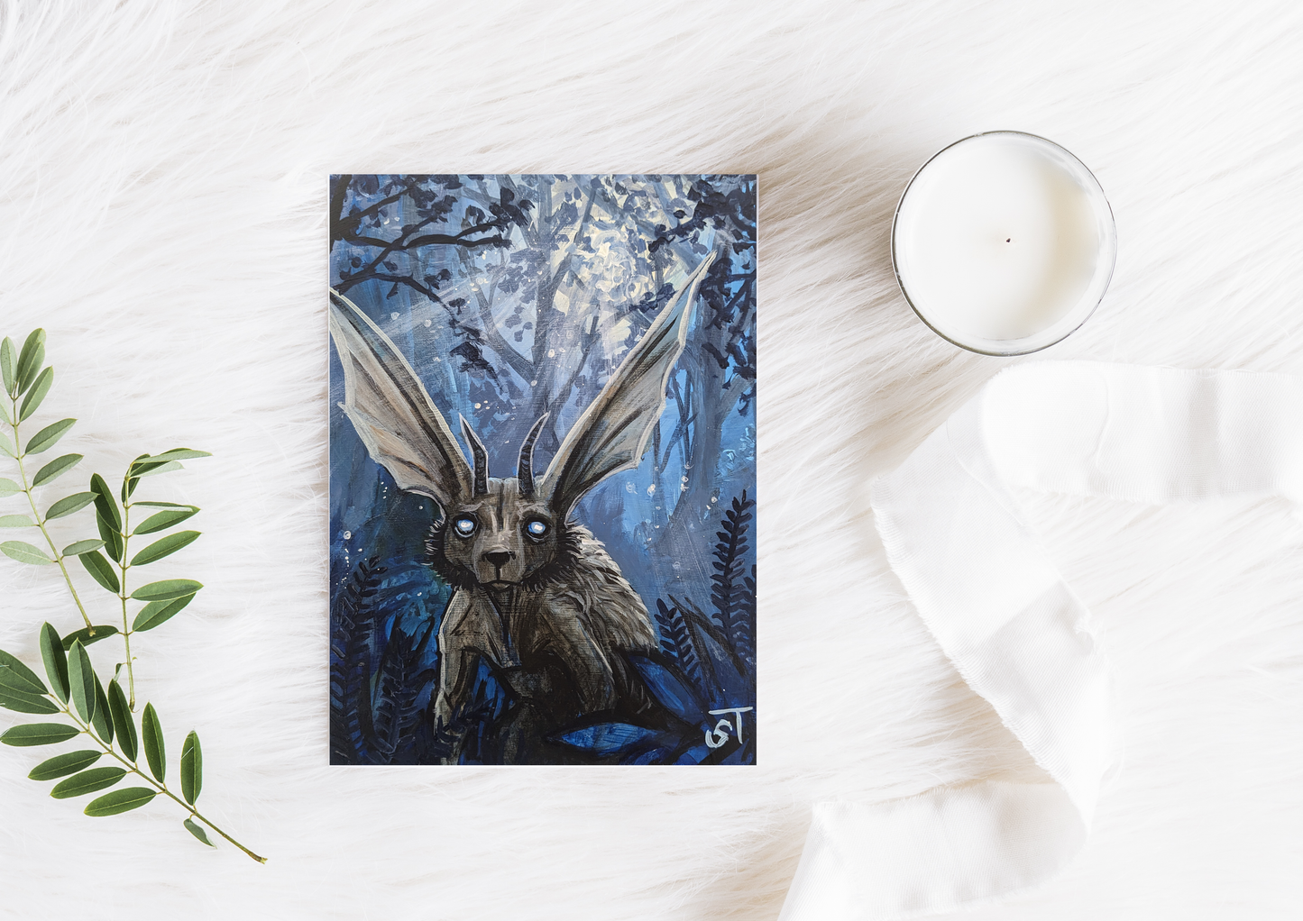 Chupacabra illustration with glowing eyes in a dark woodland scene, shown as a greeting card styled on a white surface with candle and greenery.