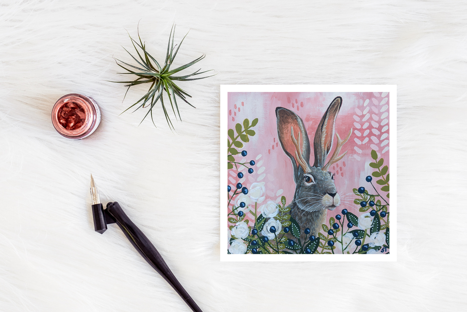 Jackalope illustration with antlers among flowers and berries on a pink background, shown as a greeting card styled on a white surface with art tools and greenery.