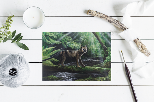 Thylacine standing near a forest stream, painted with soft light and lush greenery, shown as a greeting card styled on a white surface with yarn and a paintbrush.