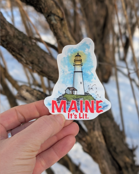 Hand holding a contour cut lighthouse vinyl sticker with the text “Maine it’s lit” outdoors among trees.