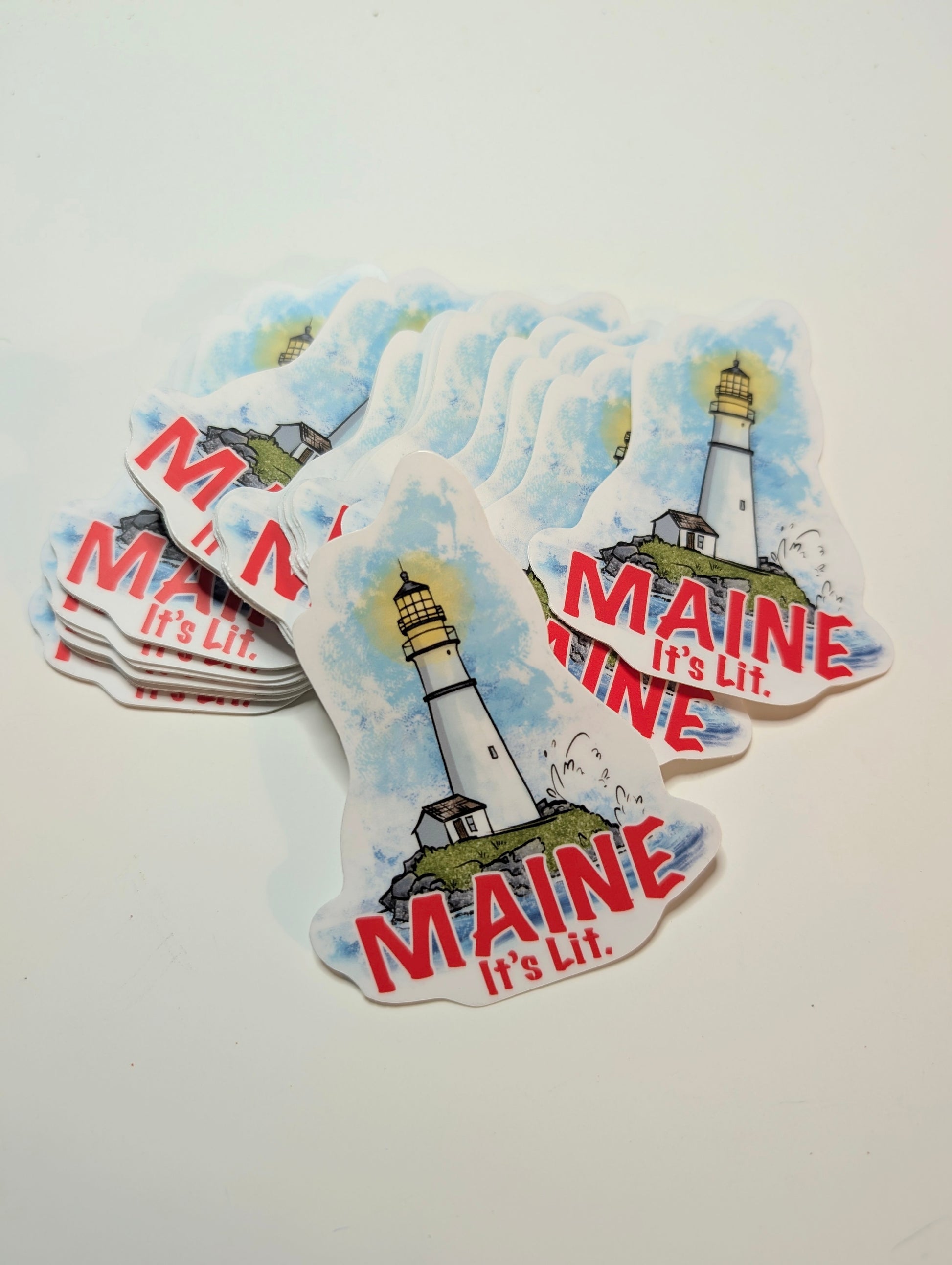Stack of contour cut lighthouse vinyl stickers with red “Maine it’s lit” text.