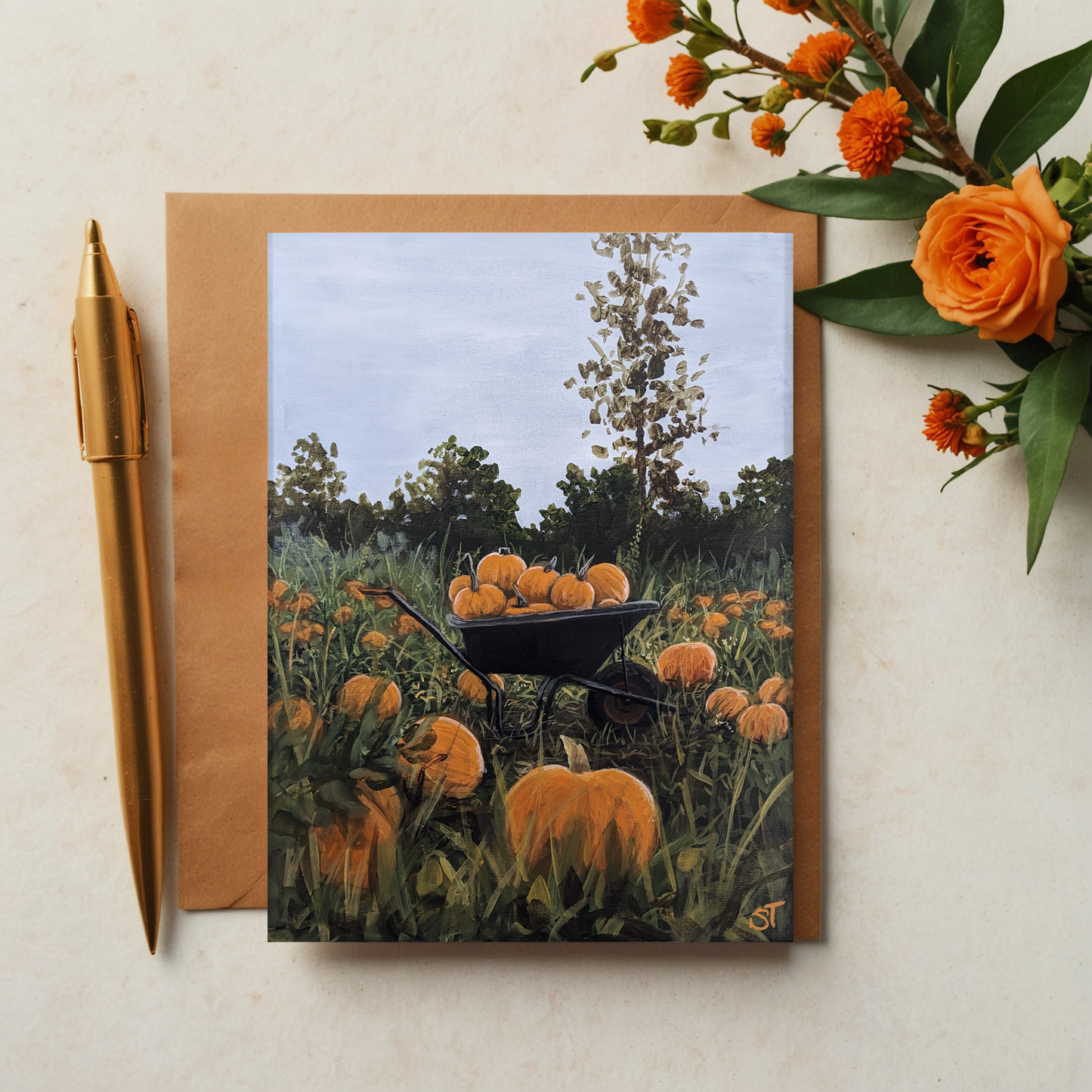 A greeting card featuring an artistic depiction of a rustic wheelbarrow filled with pumpkins, set against a background of a pumpkin patch. The card is accompanied by an envelope and a pen, positioned on a table with some flowers adding a decorative touch.