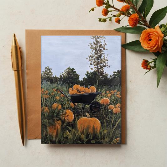 A greeting card featuring an artistic depiction of a rustic wheelbarrow filled with pumpkins, set against a background of a pumpkin patch. The card is accompanied by an envelope and a pen, positioned on a table with some flowers adding a decorative touch.