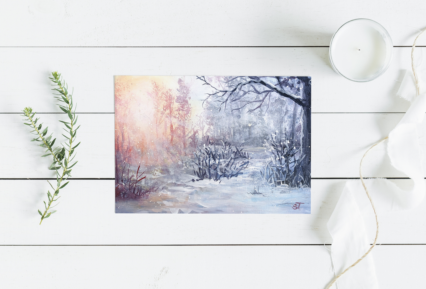 A greeting card featuring an acrylic painting of a serene winter forest scene with snow-covered trees and a warm sunset, placed on a white wooden surface with a candle and decorative foliage in the background.