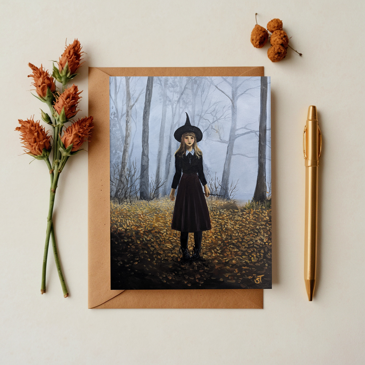 A greeting card featuring an artistic depiction of a witch girl in a forest setting, accompanied by a matching envelope and a gold pen, with decorative flowers adding a touch of autumn charm.