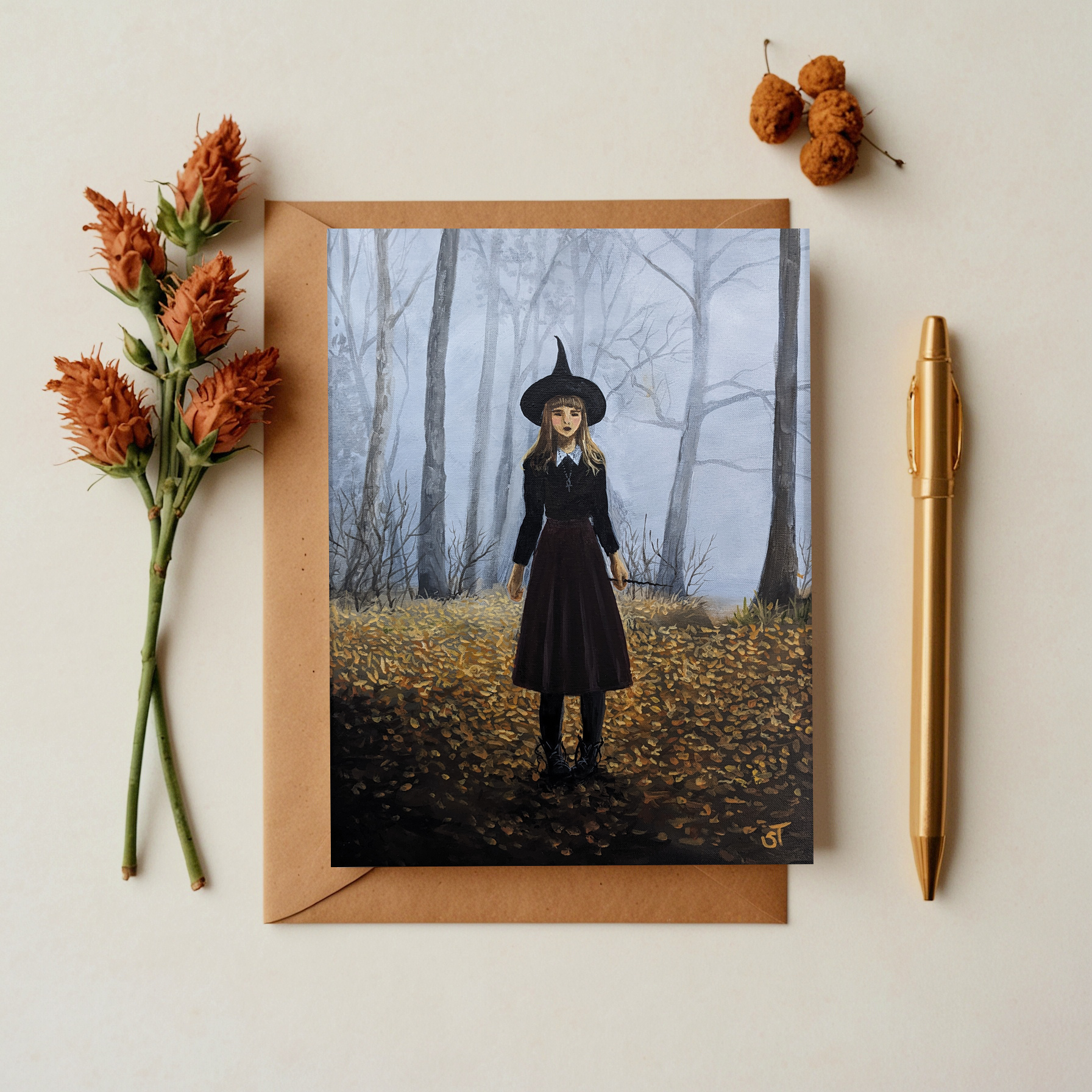 A greeting card featuring an artistic depiction of a witch girl in a forest setting, accompanied by a matching envelope and a gold pen, with decorative flowers adding a touch of autumn charm.