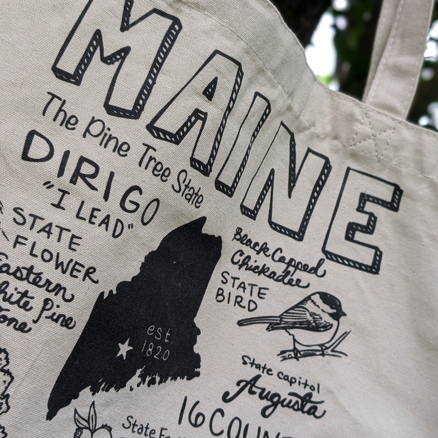 Close-up detail of the Maine-themed tote bag showing illustrated text, the state outline, and chickadee artwork.