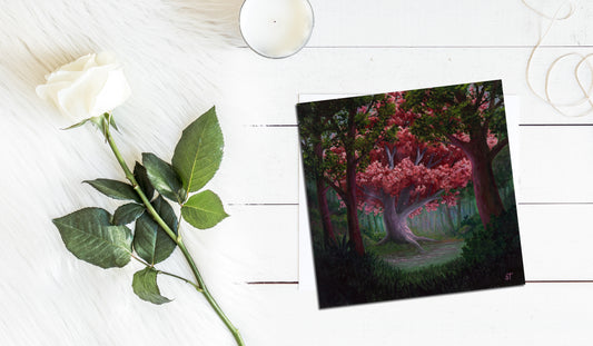 Square greeting card showing a glowing pink-leaved tree in a forest clearing, styled on a white surface with a white rose and a small candle nearby.