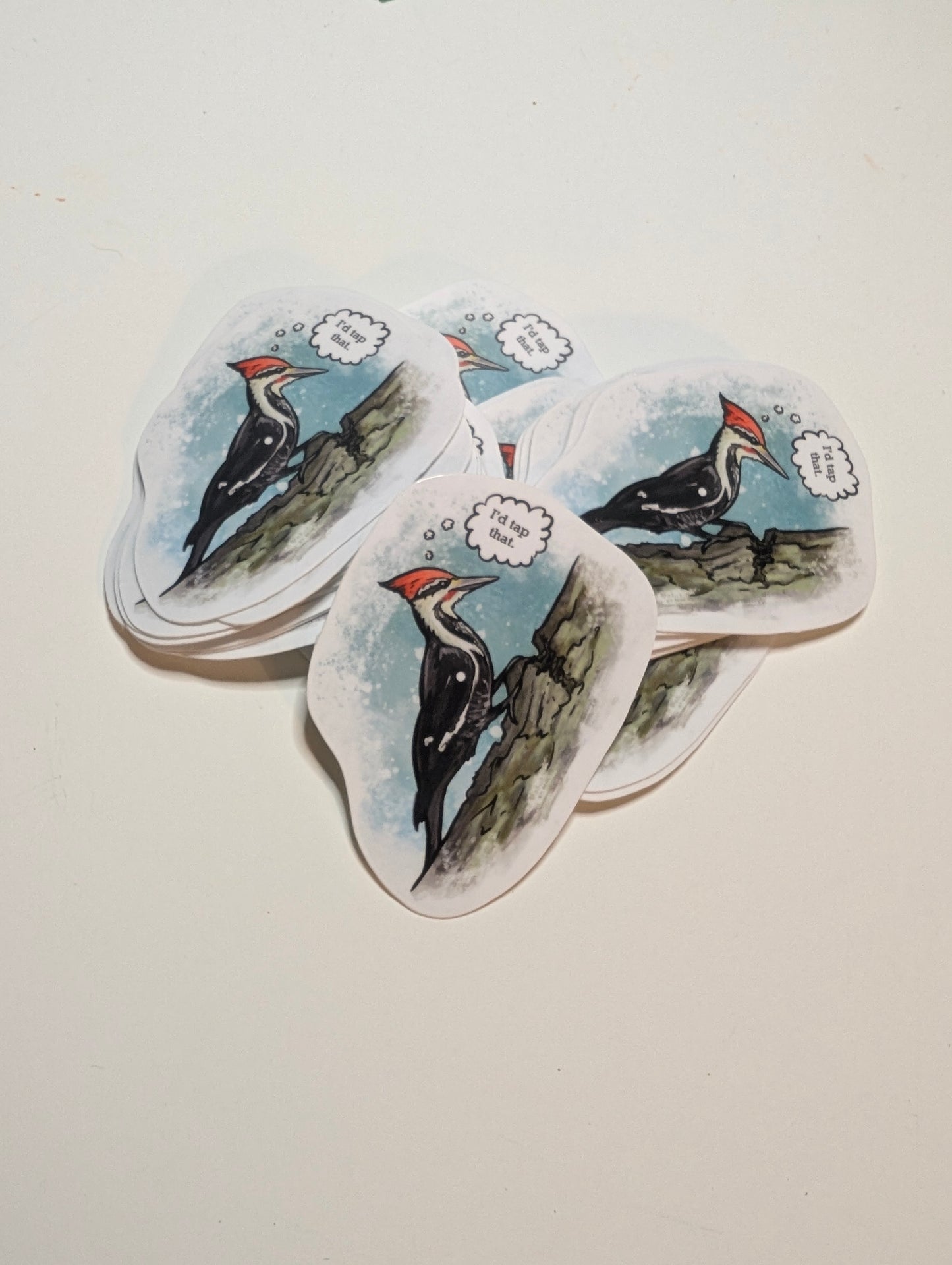 Stack of vinyl stickers featuring a pileated woodpecker on a tree trunk with a speech bubble reading “I’d tap that.”