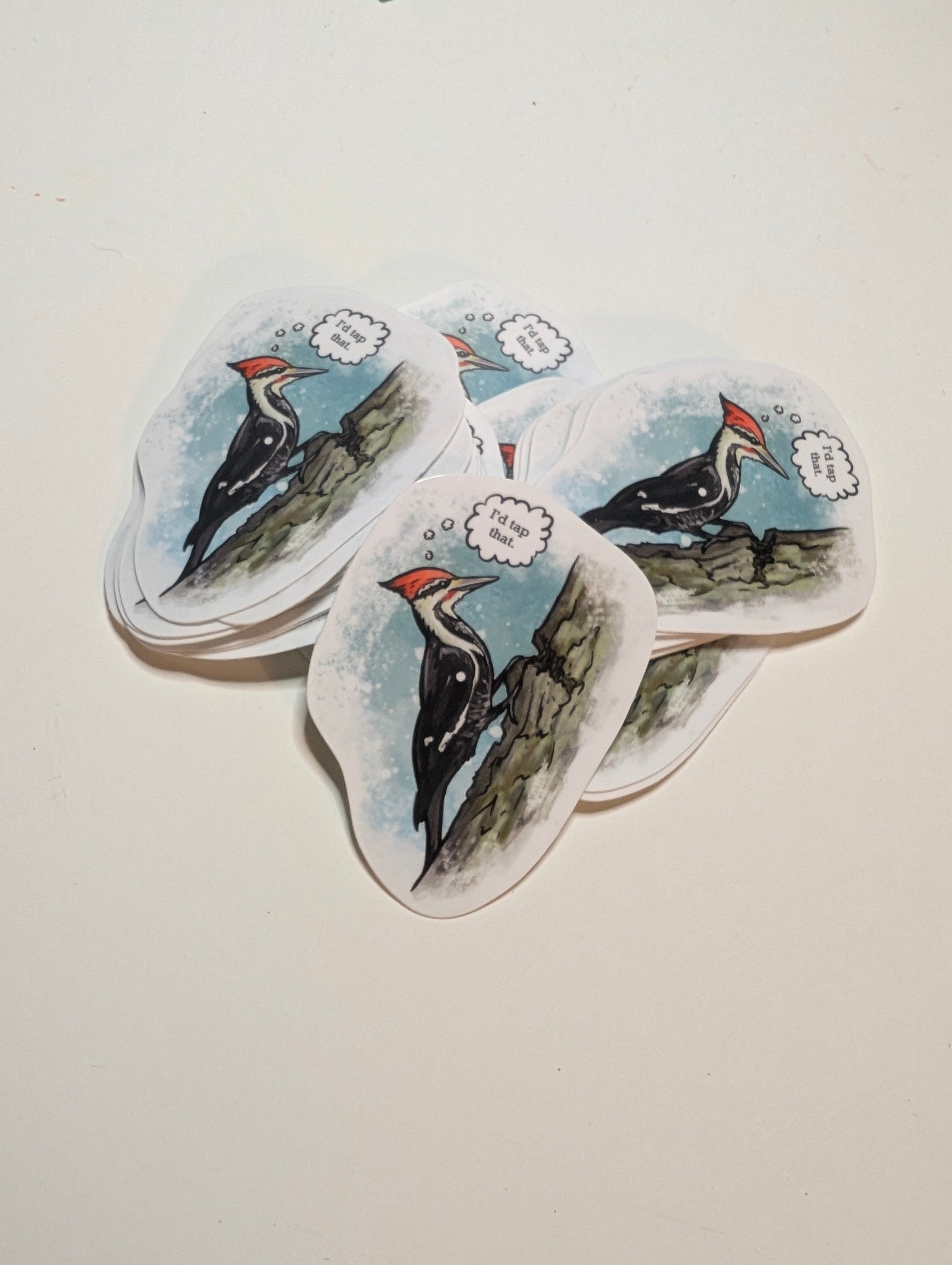 Stack of vinyl stickers featuring a pileated woodpecker on a tree trunk with a speech bubble reading “I’d tap that.”