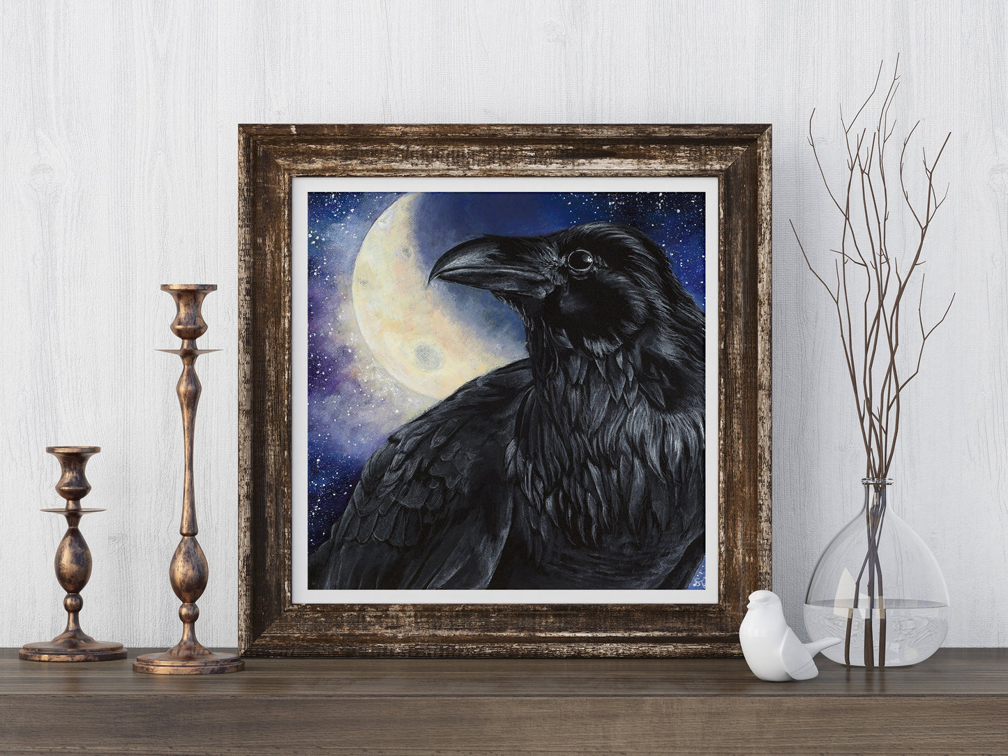 Framed art print of a black raven before a glowing full moon, styled on a wooden surface with brass candlesticks and a glass vase of bare branches.