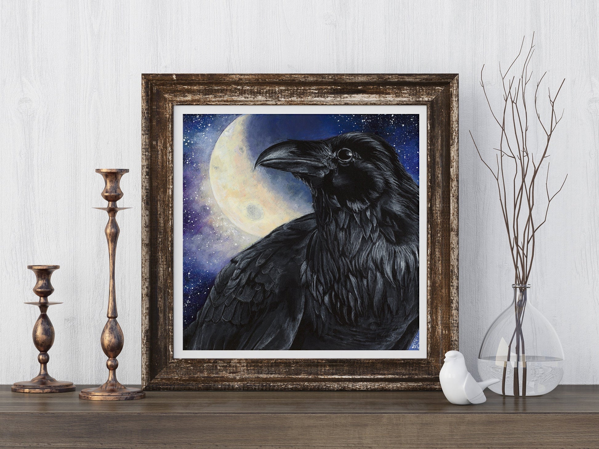 Framed art print of a black raven before a glowing full moon, styled on a wooden surface with brass candlesticks and a glass vase of bare branches.