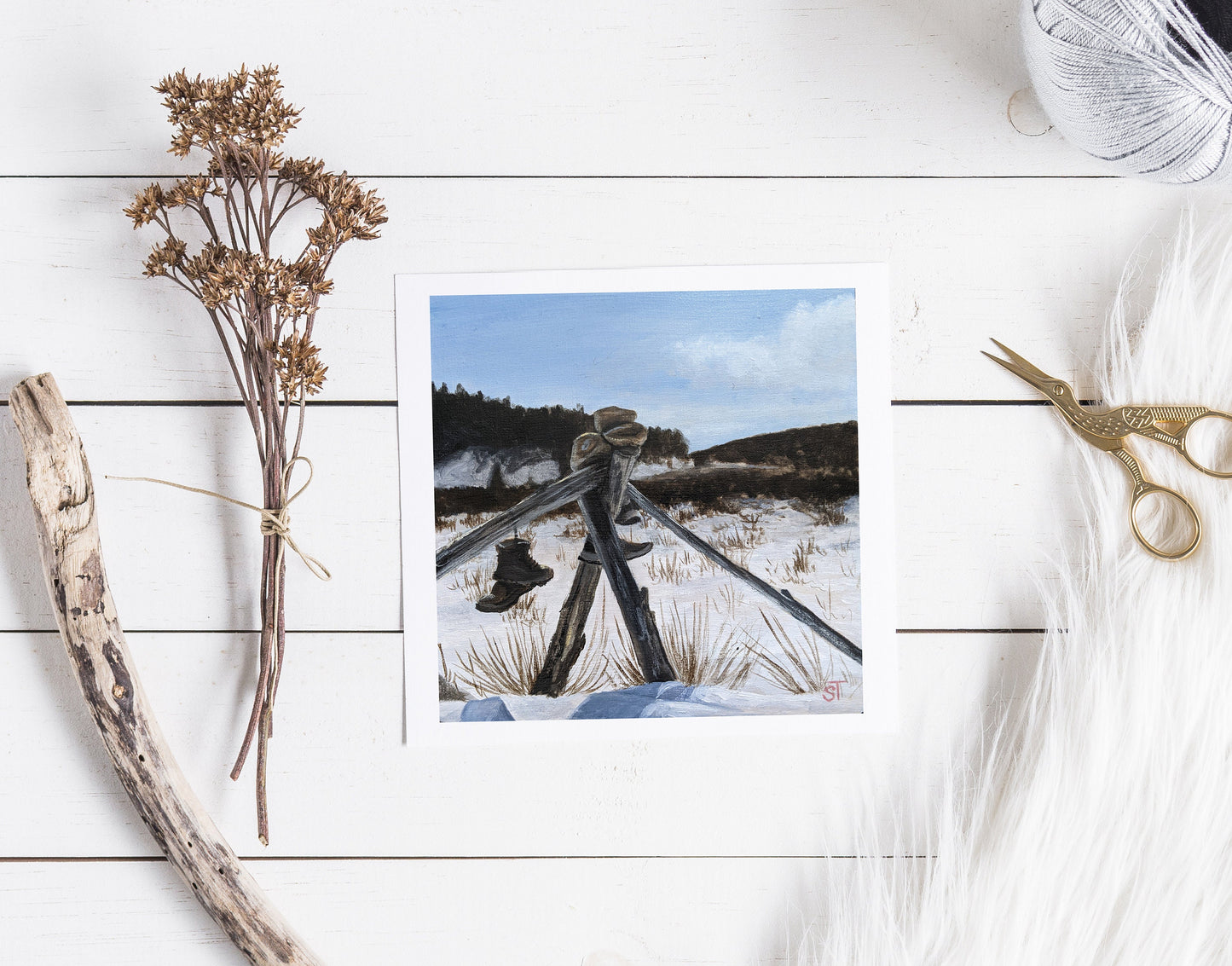 A greeting card featuring an acrylic painting of a fence in a snowy landscape, placed on a white wooden surface with decorative elements like a branch and scissors.